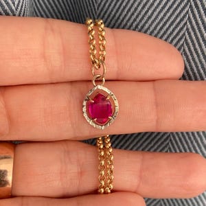 May include: A gold necklace with a pendant featuring a vibrant red gemstone. The pendant is set in a textured, silver-colored frame. The chain is a delicate, double-strand design, showcasing a classic and elegant jewelry piece.