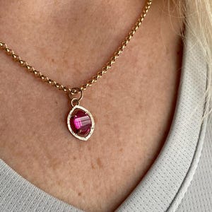 May include: Gold-toned necklace with a pendant featuring a pink gemstone. The pendant has a textured, oval-shaped frame. The chain is a simple, linked design. The necklace is worn against a light gray shirt.