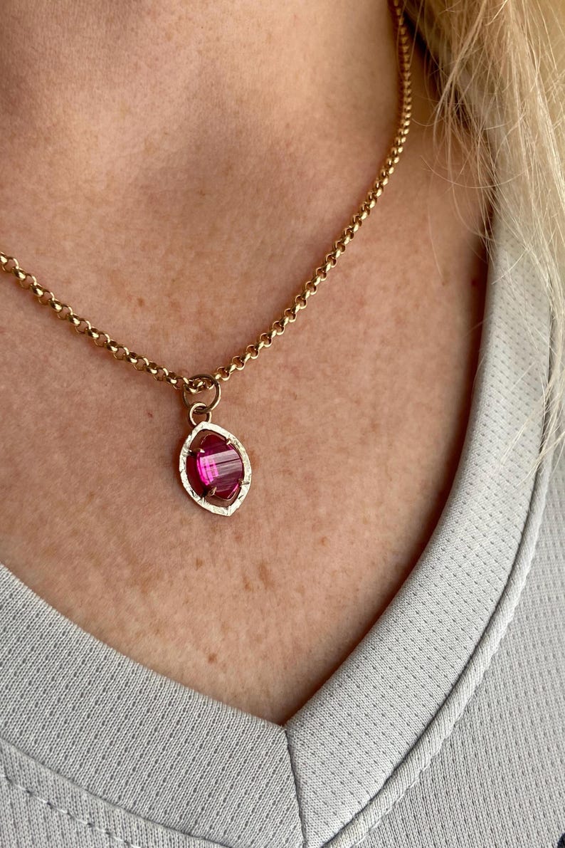 May include: Gold-toned necklace with a pendant featuring a pink gemstone. The pendant has a textured, oval-shaped frame. The chain is a simple, linked design. The necklace is worn against a light gray shirt.