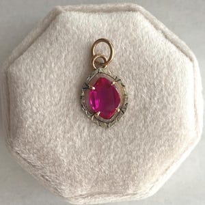 May include: A gold-toned pendant with a large, oval-shaped, magenta-colored gemstone. The gemstone is set within a detailed, silver-toned frame. The pendant is displayed on a soft, off-white surface.
