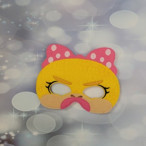 Può includere: Una maschera di feltro giallo con un'espressione arrabbiata, con un fiocco rosa a pois bianchi. La maschera ha ciglia nere e un becco rosa. Lo sfondo è azzurro con effetto bokeh.