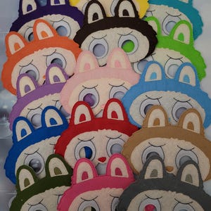 Puede incluir: Una colección de máscaras de conejito de fieltro de colores. Cada máscara presenta una cara blanca con grandes aberturas para los ojos, una nariz pequeña y una boca hacia abajo. Las máscaras vienen en varios colores, incluyendo morado, naranja, azul, verde, rosa, rojo, marrón y gris.