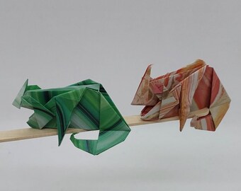 Może przedstawiać: Dwa zwierzęta origami, zielony kameleon i różowo-pomarańczowy kameleon, są zrównoważone na drewnianym patyku.