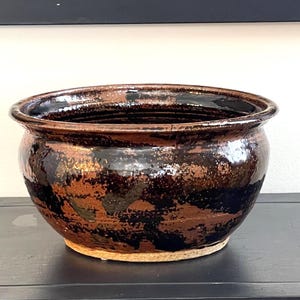 May include: A handcrafted ceramic bowl with a wide, rounded shape. The bowl features a glossy, dark brown glaze with black and copper-colored accents. The rim is slightly flared, and the base is unglazed.