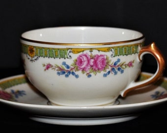May include: A white teacup and saucer with a floral design. The cup has a gold rim and a green and gold band around the top. The saucer has a similar design.
