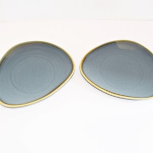 May include: Two blue-grey, organically shaped plates with a gold-colored rim. The plates are set against a white background, showcasing their unique form and color contrast. These plates are ideal for serving appetizers or small dishes.