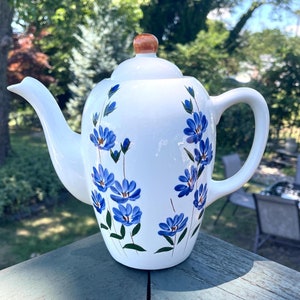 May include: White ceramic teapot with a brown lid and blue floral design. The teapot has a spout and a handle.