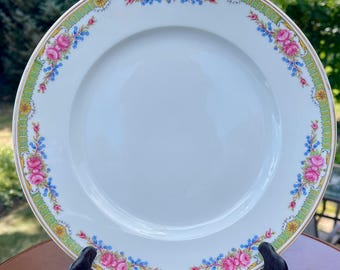 May include: White ceramic plate with a floral design. The plate features a border of pink roses, blue accents, and a green and gold trim. The plate is displayed on a black stand, showcasing its decorative details and elegant design.