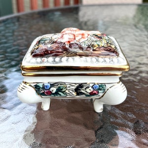 May include: A white porcelain trinket box with a gold trim and floral designs. The box has four legs and a hinged lid. The lid features a painted scene of a bird and flowers.