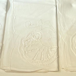 May include: A set of four rectangular white cloth napkins, each featuring a scalloped edge and a detailed tone-on-tone embroidered shell motif. The embroidery is positioned on the lower portion of each folded napkin, showcasing intricate stitching.