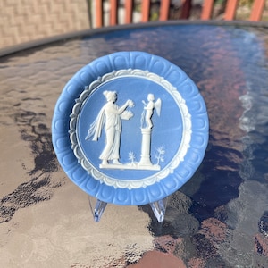May include: A blue and white ceramic plate with a classical relief design. The plate features a woman and an angel, with a decorative border. The plate is displayed on a clear plastic stand.