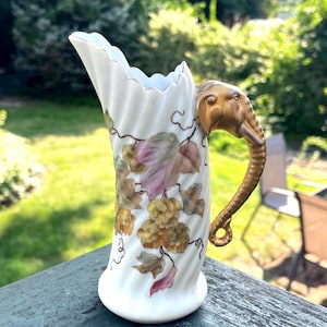 May include: White porcelain pitcher with a gold elephant head handle. The pitcher is decorated with painted grape leaves and clusters in shades of green, purple, and brown. The rim has a scalloped edge.