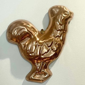 May include: A copper-colored rooster-shaped mold. The mold has detailed features, including a comb, wattle, and tail feathers. The rooster is a kitchenware item, likely used for baking or decorative purposes. The mold is made of a shiny, reflective metal.