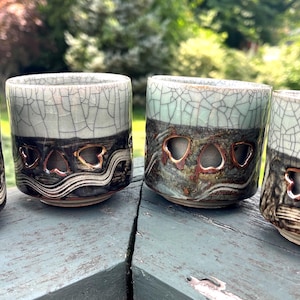 May include: Four ceramic cups with a unique design. The cups have a light gray, crackled glaze on the top, transitioning to a dark band with heart-shaped cutouts and wavy lines. The cups are on a weathered wooden surface.