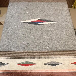 May include: A rectangular woven table runner featuring a speckled gray base with wide cream-colored bands at both ends. The ends are decorated with repeating geometric motifs in red, green, and black, accented by thin brown and orange stripes.