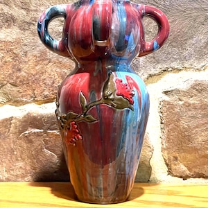 May include: A ceramic vase with a unique shape and two handles. The vase features a blend of red, blue, and green vertical stripes. A floral design with red berries and gold accents adorns the side. The vase is a decorative item.