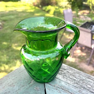 May include: A small, green glass pitcher with a handle and a spout. The pitcher has a textured diamond pattern and a black rim. The pitcher is sitting on a weathered wooden surface, with a blurred green background.