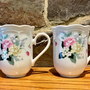 May include: Two white ceramic mugs with floral designs. Each mug features a scalloped rim and a curved handle. The floral pattern includes pink, yellow, and green flowers with small red berries. The mugs are set on a wooden surface.