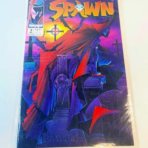 May include: A Spawn comic book issue number 2, published by Image Comics in June. The cover features a dark, caped figure perched on a tombstone in a graveyard at sunset, with the title Spawn in yellow and the name Simmons at the bottom.
