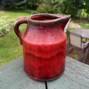 May include: A handmade ceramic pitcher with a red glaze, featuring a dark brown rim and handle. The pitcher has a classic design with a spout for easy pouring. The exterior has a textured appearance, adding to its rustic charm.