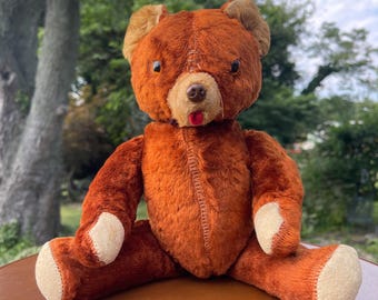 May include: A vintage teddy bear with brown fur, tan ears, and a tan snout. The bear has a red tongue and white paw pads. It is sitting on a wooden surface with a blurred green background.