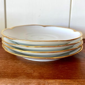 May include: A stack of four white ceramic plates with scalloped edges and gold rims. The plates are stacked on top of each other, showcasing the elegant design. The plates are on a wooden surface.