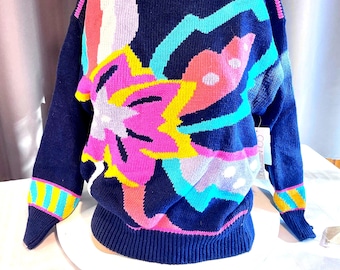 May include: Navy blue sweater with a vibrant floral design in pink, yellow, blue, and white. The sweater features a crew neck and ribbed cuffs with colorful stripes. A tag is attached to the sweater.