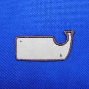 May include: A white whale-shaped patch with a brown outline is centered on a blue fabric background. The patch has a small dot and a tiny symbol. The fabric has a textured appearance.