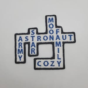 May include: Embroidered patch in a crossword puzzle design. The patch features the words "ARMY," "ASTRONAUT," "MOON," "FAMILY," and "COZY" in blue lettering on a white background, outlined in black.