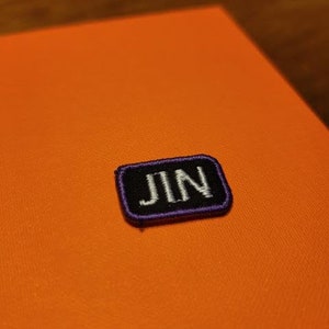 May include: A small, rectangular patch with the name "JIN" embroidered in white on a black background. The patch has a purple border and is set against an orange background.