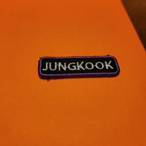 May include: A rectangular patch with the name "JUNGKOOK" embroidered in white on a black background, outlined with purple stitching. The patch is placed on an orange surface.
