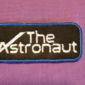 May include: A black and blue embroidered patch with white text that reads "The Astronaut".