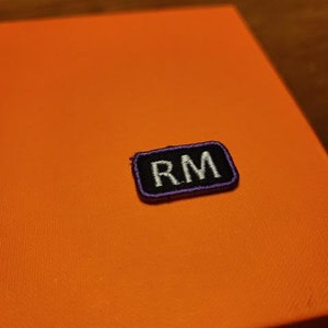 May include: An orange box with a small, rectangular patch. The patch is black with white embroidered letters "RM" and a purple border. The box has a textured surface.