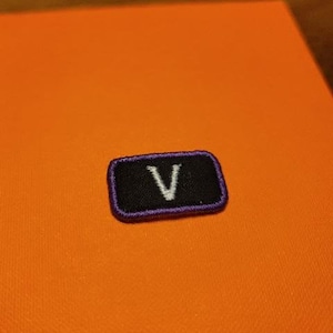 May include: A rectangular black patch with a purple border and a white "V" in the center. The patch is set against an orange background.