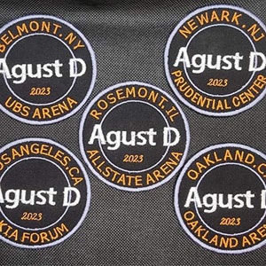 May include: Five black and white circular patches with the text "Agust D" in white and the year "2023" in orange. Each patch has a different venue name in orange text around the outside edge. The venues are: UBS Arena, Prudential Center, Allstate Arena, Kia Forum, and Oakland Arena.