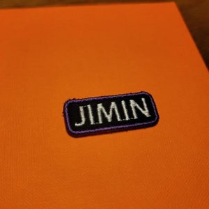 May include: A black and white embroidered patch with purple stitching. The patch says "JIMIN" in white letters.