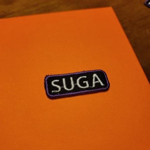May include: An orange surface with a rectangular patch that reads "SUGA" in white letters. The patch has a black border with purple stitching. The background is a solid orange color.