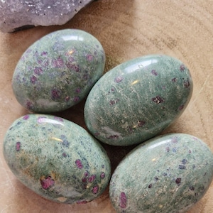 May include: Four polished, oval-shaped stones with a green base colour and red inclusions. The stones are smooth and reflective, with a natural, organic appearance. A piece of purple crystal is in the background.