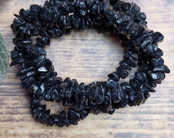 May include: A black gemstone bracelet composed of small, irregularly shaped beads. The bracelet is displayed on a wooden surface, with a sprig of greenery in the background. The beads have a glossy finish.