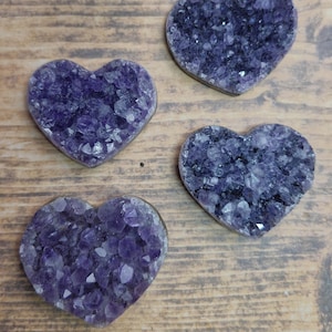 May include: Four heart-shaped amethyst crystal geodes. The geodes are a deep purple color with visible crystalline structures. They are arranged on a wooden surface, showcasing their natural beauty.