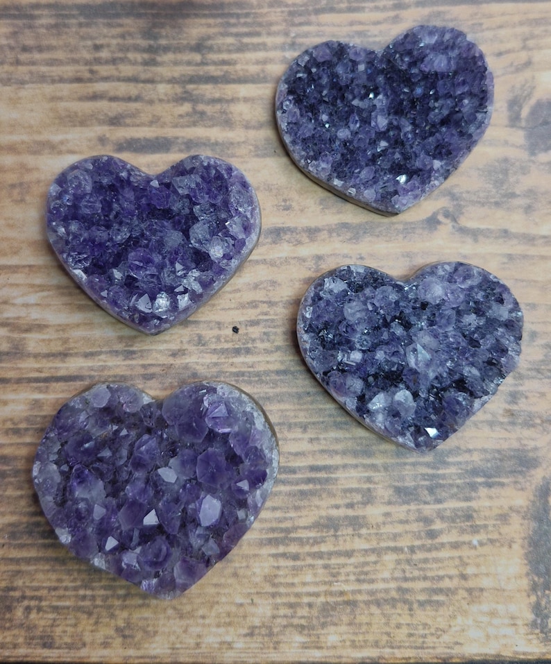 May include: Four heart-shaped amethyst crystal geodes. The geodes are a deep purple color with visible crystalline structures. They are arranged on a wooden surface, showcasing their natural beauty.