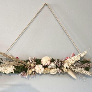 May include: A decorative hanging floral display with dried flowers, grasses, and a seashell. The arrangement is set on a green moss base and suspended by a natural rope. The color scheme includes beige, white, and red accents.
