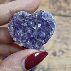 May include: A heart-shaped amethyst crystal, showcasing a deep purple hue. The crystal's surface is composed of numerous small, sparkling facets, creating a textured appearance. The heart is held against a blurred wooden background.