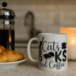 Peut inclure: Une tasse blanche avec l'inscription "Cats Books And Coffee" en noir. Deux croissants sont posés sur une assiette blanche. Une cafetière à piston et une bougie allumée sont en arrière-plan.