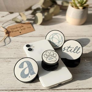 Puede incluir: Un teléfono inteligente blanco con varios agarres redondos para teléfono. Los agarres presentan varios diseños, incluyendo la letra azul "a", una huella de pata con la palabra "Love", un patrón geométrico y la palabra "still". Una pequeña planta suculenta en maceta está en el fondo.