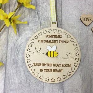 May include: A wooden round ornament with a bee design and the text "Sometimes the smallest things take up the most room in your heart". The ornament is surrounded by a border of small hearts. The ornament is hanging from a yellow and white ribbon.