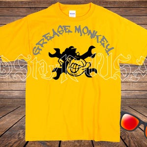 May include: Yellow t-shirt with the text "GREASE MONKEY" arched above a black graphic of a monkey with crossed wrenches. The monkey is smiling and has a wrench in its mouth. Sunglasses with red lenses are in the bottom right corner.