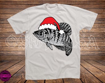 Puede incluir: Camiseta blanca con un diseño detallado de pez negro y gris que lleva un gorro de Papá Noel rojo con ribete blanco. La camiseta tiene cuello redondo y mangas cortas. El diseño está centrado en la parte delantera.