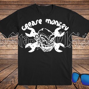 May include: Black t-shirt with a white graphic featuring a grinning monkey face, crossed wrenches, and the words "Grease Monkey." The design is bold and eye-catching. Sunglasses with blue lenses rest on a wooden surface.