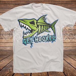 May include: White t-shirt featuring a green and black shark skeleton graphic with the text "GET HOOKED" in blue and white. The design has a cartoonish style, with a distressed look. The shirt is made of a soft material.